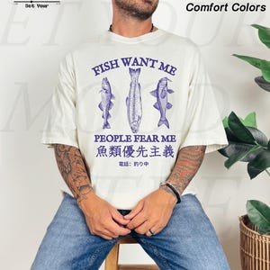 May include: Ivory-colored t-shirt with the text "FISH WANT ME PEOPLE FEAR ME" and Japanese text. The shirt features three blue fish illustrations. The words "Comfort Colors" are also visible.