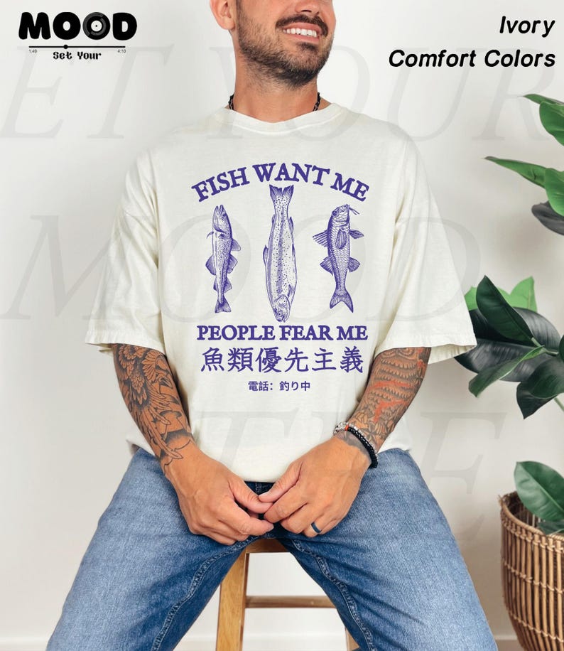 May include: Ivory-colored t-shirt with the text "FISH WANT ME PEOPLE FEAR ME" and Japanese text. The shirt features three blue fish illustrations. The words "Comfort Colors" are also visible.