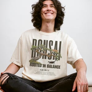 May include: Ivory-colored t-shirt with a bonsai tree graphic and the text "BONSAI ROOTED IN BALANCE." The shirt also features Japanese text and the words "Ivory Comfort Colors." The person is wearing black jeans.