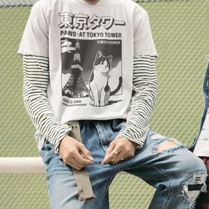 以下が含まれることがあります： 猫のグラフィックと「PAWS AT TOKYO TOWER」の文字が入った白いTシャツを着た人が、ストライプの長袖シャツの上に、破れた青いジーンズと黒いブーツを合わせています。シャツには日本語のテキストも入っています。