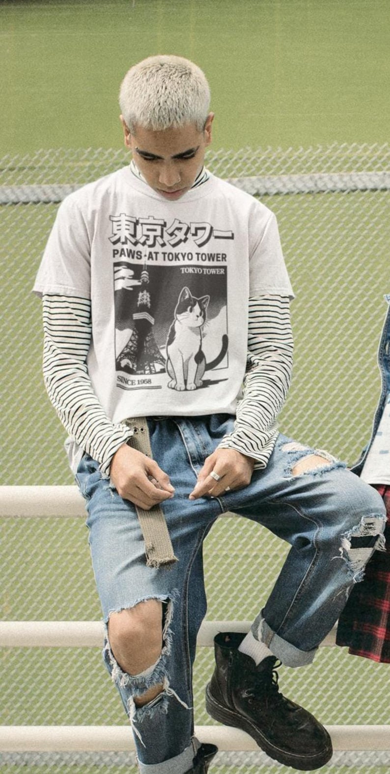 以下が含まれることがあります： 猫のグラフィックと「PAWS AT TOKYO TOWER」の文字が入った白いTシャツを着た人が、ストライプの長袖シャツの上に、破れた青いジーンズと黒いブーツを合わせています。シャツには日本語のテキストも入っています。