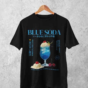 May include: Black t-shirt featuring a blue soda graphic with Japanese text. The design includes a blue cocktail with whipped cream and cherries, and the words "BLUE SODA" in large blue letters.
