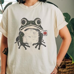 May include: Cream-colored t-shirt featuring a large, stylized frog illustration in shades of gray and black. The frog has large eyes and is centered on the shirt. A small red and white graphic is to the right of the frog.