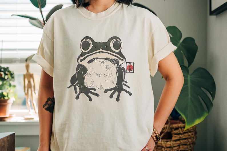 May include: Cream-colored t-shirt featuring a large, stylized frog illustration in shades of gray and black. The frog has large eyes and is centered on the shirt. A small red and white graphic is to the right of the frog.