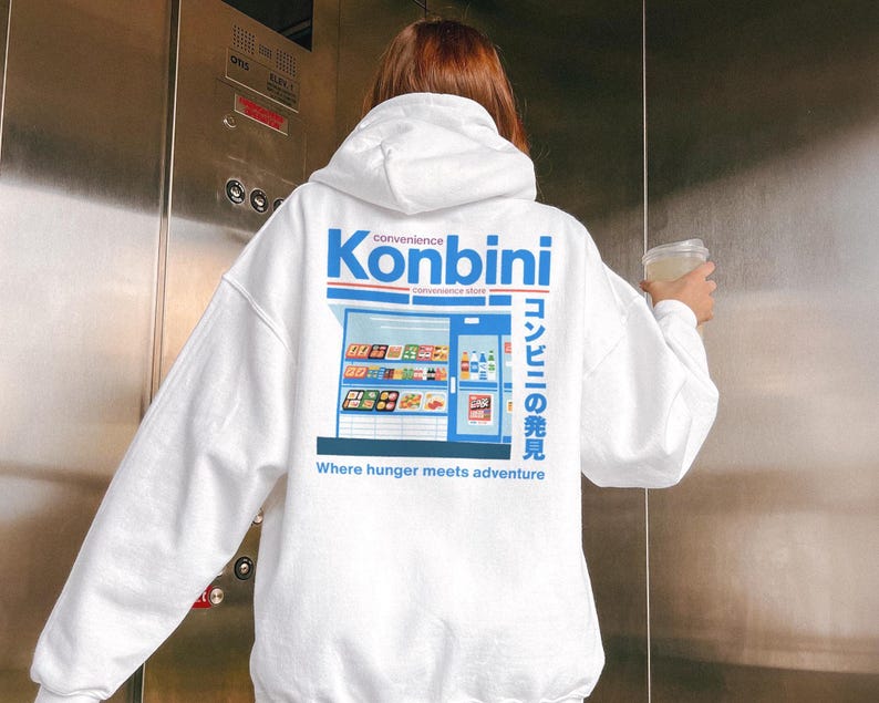 White hoodie featuring a blue Japanese konbini (convenience store) graphic on the back. The design includes Japanese text and the phrase “Where hunger meets adventure.”