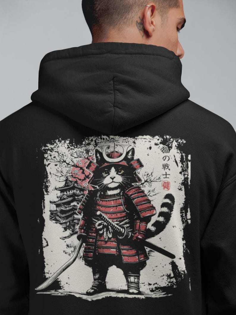 Back view of a black hoodie featuring a detailed Japanese-style illustration of a cat dressed as a samurai warrior. The cat wears red armor, holds a katana, and stands in front of a traditional pagoda with cherry blossoms. The artwork has a distressed ink texture with Japanese kanji text on the right side, giving it a traditional Ukiyo-e and manga-inspired aesthetic.