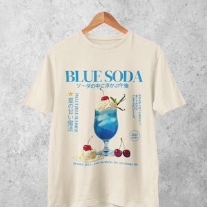 May include: Cream-colored t-shirt with a blue graphic of a blue soda drink with cherries and whipped cream. Text includes "BLUE SODA" and Japanese characters. The phrase "Bubbly, blue, and avoiding all my problems" is also present.