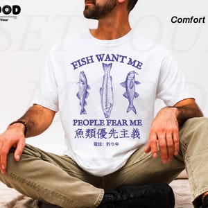 May include: White t-shirt with a blue graphic of three fish and the text "FISH WANT ME PEOPLE FEAR ME". The shirt also includes Japanese text. The shirt is made of a comfortable material.