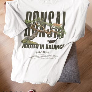 May include: White t-shirt with a bonsai tree graphic and the text "BONSAI ROOTED IN BALANCE". The shirt also features Japanese text and the words "White Comfort Colors". The shirt is folded on a chair.