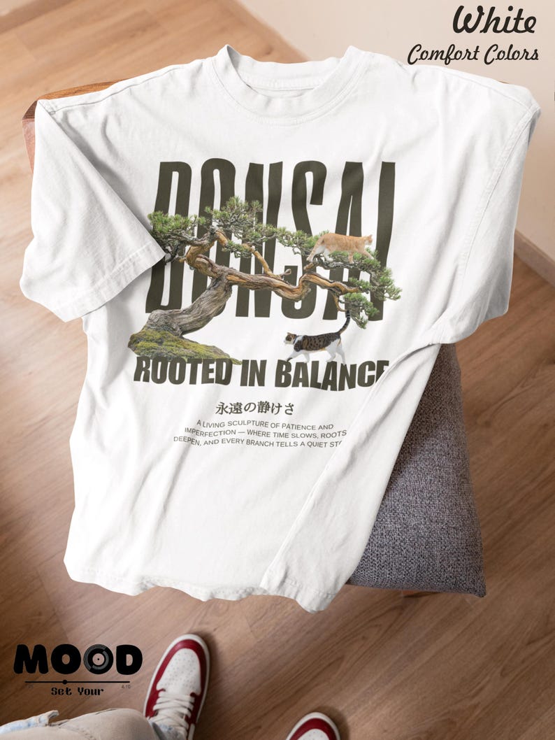 May include: White t-shirt with a bonsai tree graphic and the text "BONSAI ROOTED IN BALANCE". The shirt also features Japanese text and the words "White Comfort Colors". The shirt is folded on a chair.