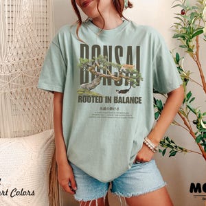 May include: A light green t-shirt with a bonsai tree graphic and the text "BONSAI" above the tree and "ROOTED IN BALANCE" below. The shirt also has Japanese text. The shirt is part of the "Bay Comfort Colors" collection.