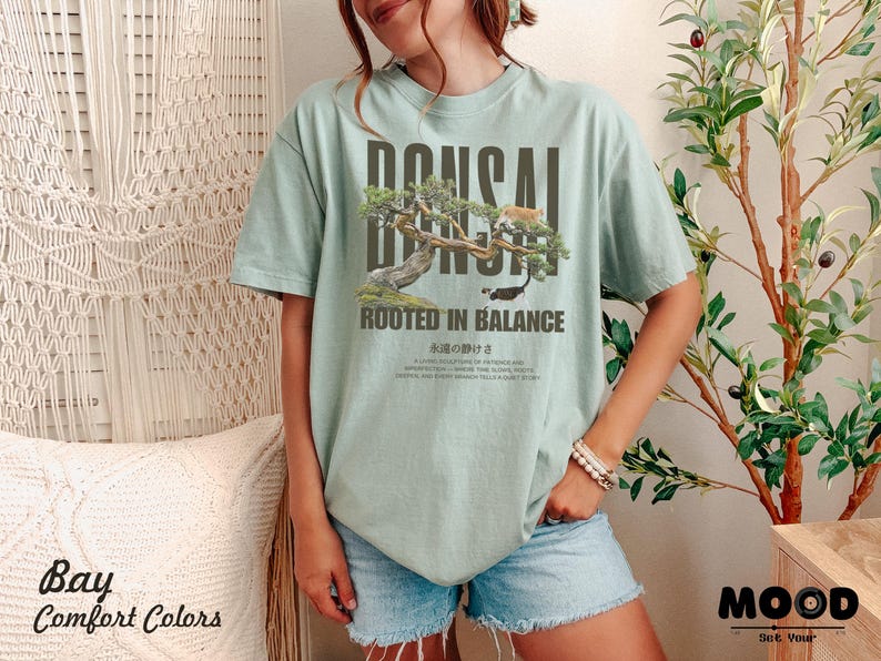 May include: A light green t-shirt with a bonsai tree graphic and the text "BONSAI" above the tree and "ROOTED IN BALANCE" below. The shirt also has Japanese text. The shirt is part of the "Bay Comfort Colors" collection.