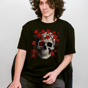 May include: Black t-shirt featuring a detailed skull graphic with red cherry blossoms and Japanese text. The skull has one red eye and is surrounded by the flowers. The shirt is a classic crew neck style.