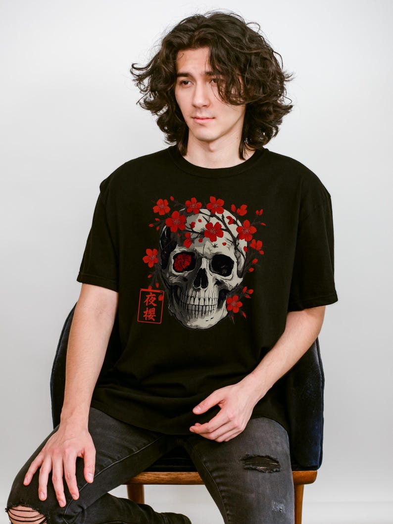 May include: Black t-shirt featuring a detailed skull graphic with red cherry blossoms and Japanese text. The skull has one red eye and is surrounded by the flowers. The shirt is a classic crew neck style.