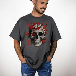 May include: Dark gray t-shirt featuring a detailed skull graphic adorned with red flowers. The skull has one red eye and is surrounded by red blossoms. The shirt has a relaxed fit and short sleeves, perfect for casual wear.