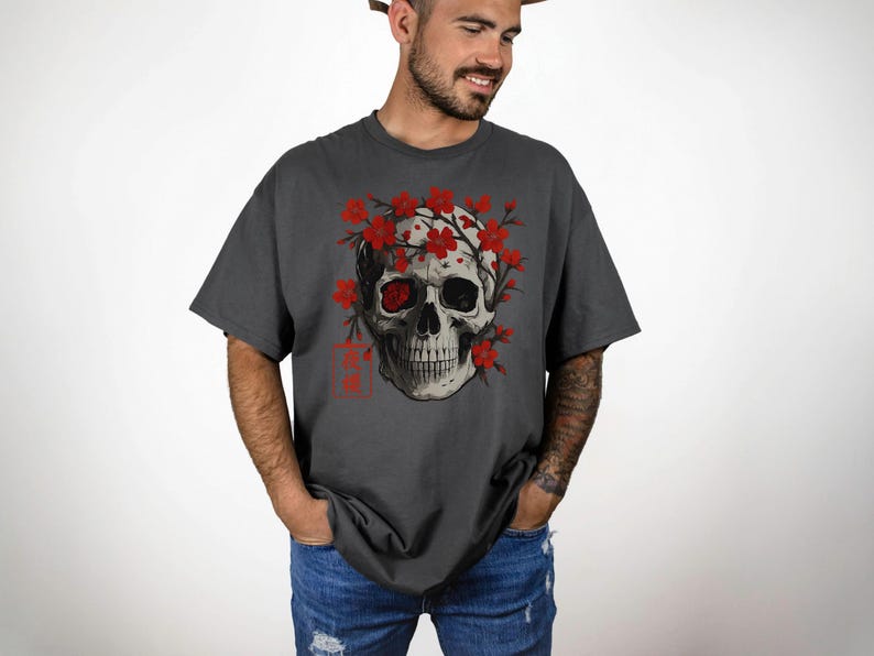 May include: Dark gray t-shirt featuring a detailed skull graphic adorned with red flowers. The skull has one red eye and is surrounded by red blossoms. The shirt has a relaxed fit and short sleeves, perfect for casual wear.