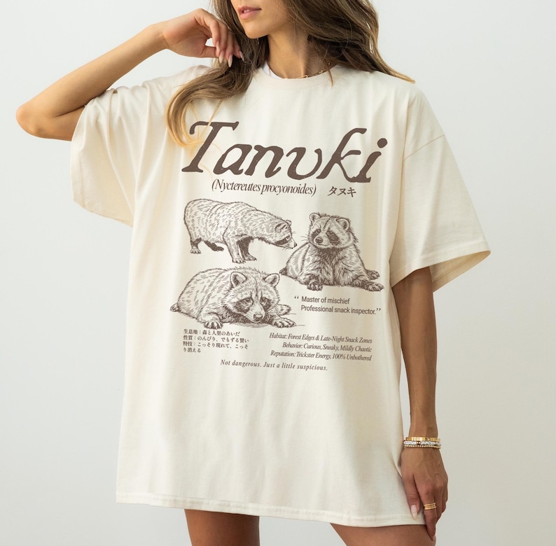 May include: Cream-colored oversized t-shirt featuring a graphic of three raccoons and the word "Tanuki" in brown. The shirt includes text in English and Japanese, with additional text describing the raccoon's habitat and behavior.