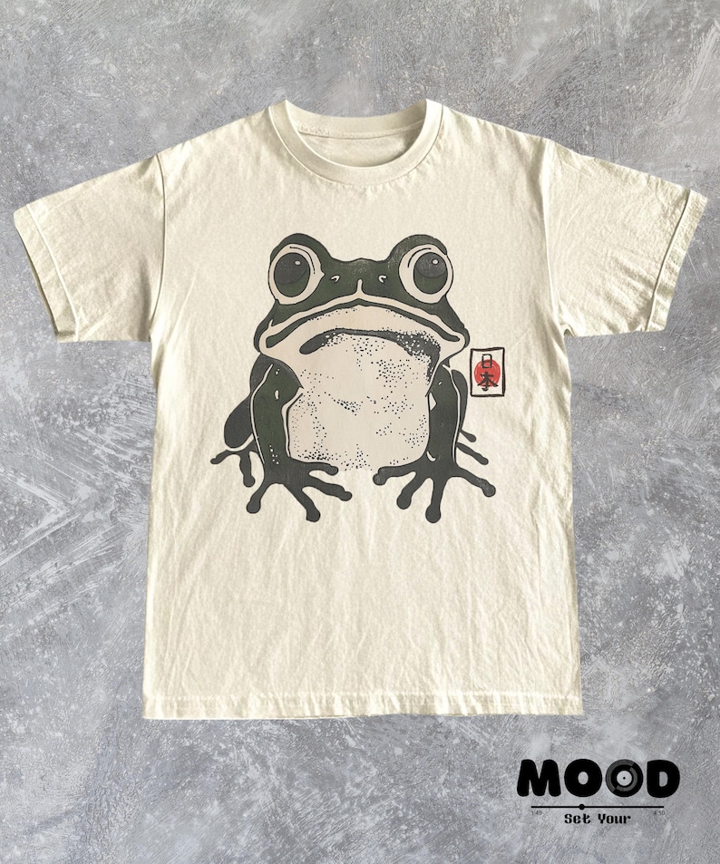 May include: Cream-colored t-shirt featuring a large, stylized frog graphic in black and green. The frog has large eyes and is centered on the shirt. A small red and white Japanese symbol is on the right side. The shirt has a crew neck.