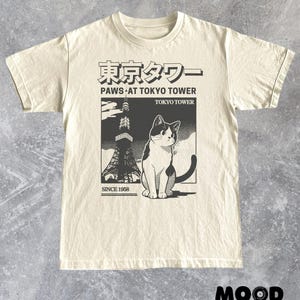 以下が含まれることがあります： 東京タワーの前にいる猫のグラフィックが特徴のクリーム色のTシャツ。デザインには日本語のテキストと「PAWS AT TOKYO TOWER TOKYO TOWER SINCE 1958」というフレーズが含まれています。Tシャツはクルーネックで半袖です。