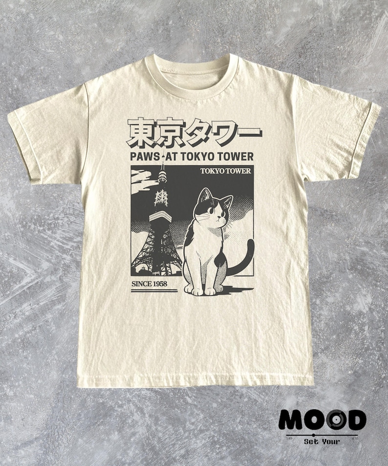 以下が含まれることがあります： 東京タワーの前にいる猫のグラフィックが特徴のクリーム色のTシャツ。デザインには日本語のテキストと「PAWS AT TOKYO TOWER TOKYO TOWER SINCE 1958」というフレーズが含まれています。Tシャツはクルーネックで半袖です。