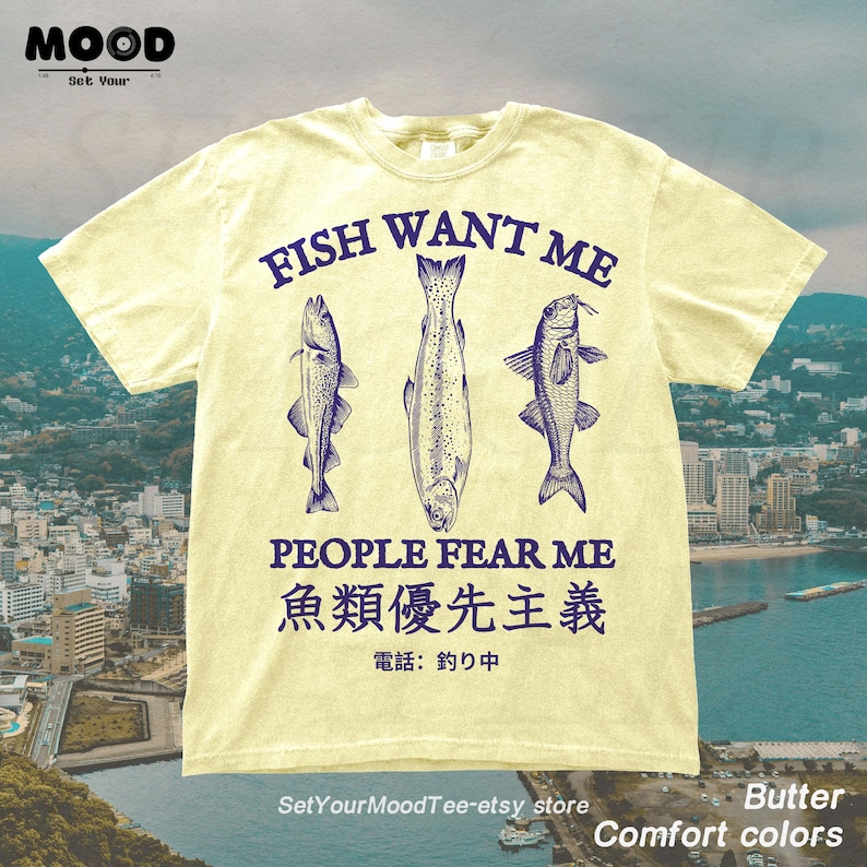 May include: Butter-colored t-shirt with the text "FISH WANT ME PEOPLE FEAR ME" and Japanese text. The shirt features three detailed fish illustrations in navy blue. The Etsy store name is visible at the bottom.