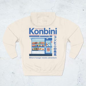 May include: Cream-colored hoodie featuring a graphic of a convenience store with the word "Konbini" in blue and Japanese text. The phrase "Where hunger meets adventure" is printed below the store illustration.