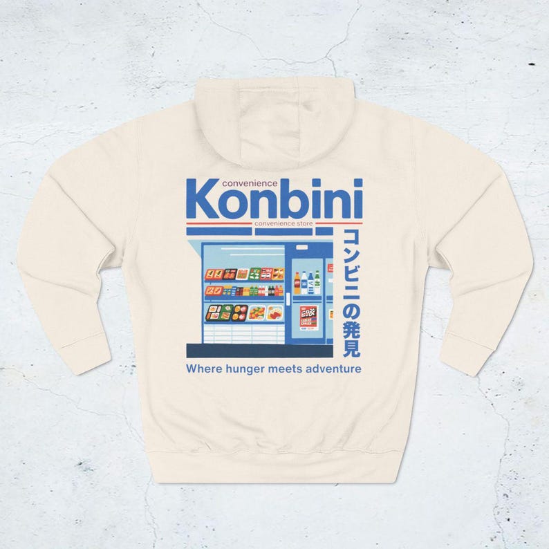 May include: Cream-colored hoodie featuring a graphic of a convenience store with the word "Konbini" in blue and Japanese text. The phrase "Where hunger meets adventure" is printed below the store illustration.