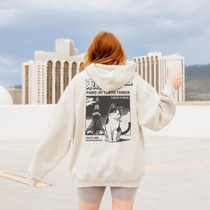 May include: A cream-coloured hoodie with a graphic on the back featuring a black and white cat, the Tokyo Tower, and the text "PAWS AT TOKYO TOWER TOKYO TOWER SINCE 1988". The hoodie has a drawstring hood and long sleeves.