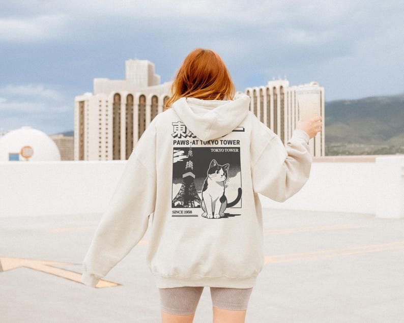 May include: A cream-coloured hoodie with a graphic on the back featuring a black and white cat, the Tokyo Tower, and the text "PAWS AT TOKYO TOWER TOKYO TOWER SINCE 1988". The hoodie has a drawstring hood and long sleeves.