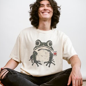 May include: Cream-colored t-shirt featuring a large, stylized frog graphic in shades of green and black. The frog design has a Japanese aesthetic. The shirt is paired with black jeans.