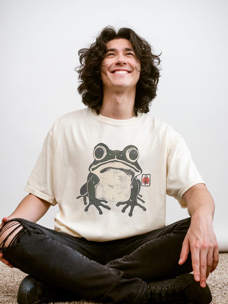 May include: Cream-colored t-shirt featuring a large, stylized frog graphic in shades of green and black. The frog design has a Japanese aesthetic. The shirt is paired with black jeans.