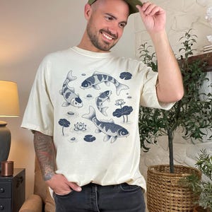 May include: Cream-colored t-shirt featuring a koi pond design. The graphic includes several koi fish, water lilies, and lily pads in shades of blue. The shirt has a relaxed fit and short sleeves.