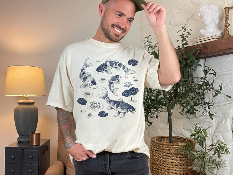 May include: Cream-colored t-shirt featuring a koi pond design. The graphic includes several koi fish, water lilies, and lily pads in shades of blue. The shirt has a relaxed fit and short sleeves.