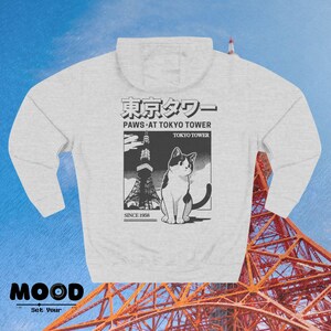 May include: Pale grey hoodie with a graphic of a cat and the Tokyo Tower. The graphic includes Japanese text and the phrase "Paws at Tokyo Tower" with "Since 1958" below. The hoodie has a hood and a front pocket.