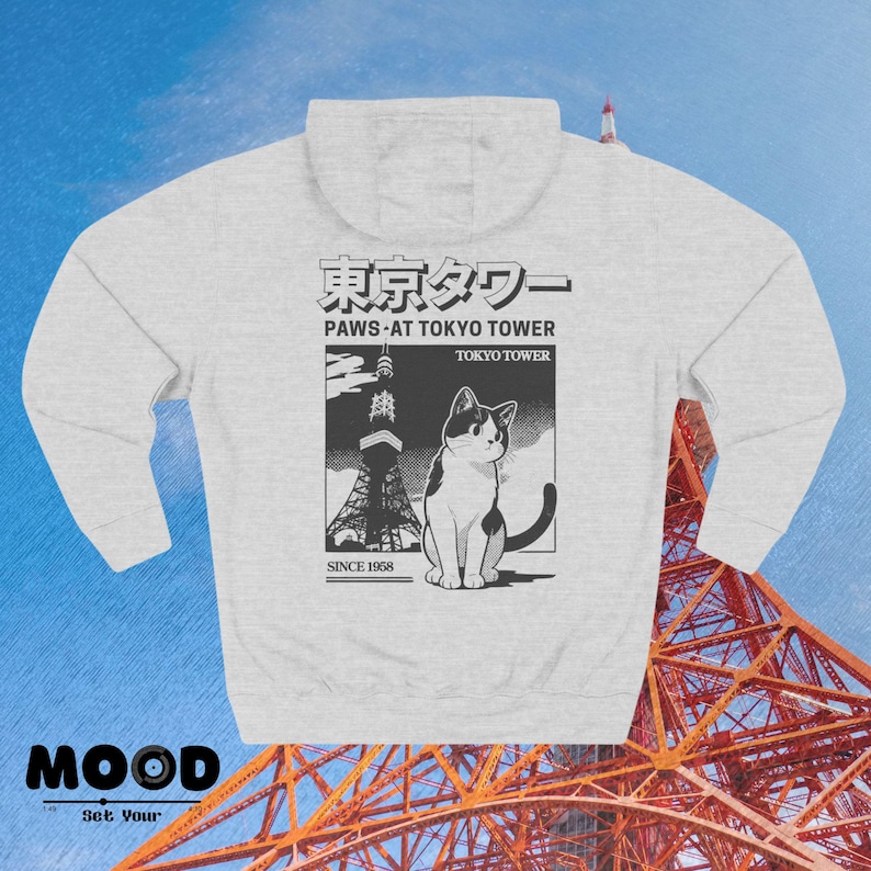 May include: Pale grey hoodie with a graphic of a cat and the Tokyo Tower. The graphic includes Japanese text and the phrase "Paws at Tokyo Tower" with "Since 1958" below. The hoodie has a hood and a front pocket.