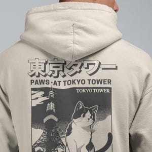 May include: Cream hoodie with a graphic of a cat and the Tokyo Tower. The design includes Japanese text and the phrase "Paws at Tokyo Tower" with "Since 1958" below the image. The cat is black and white.