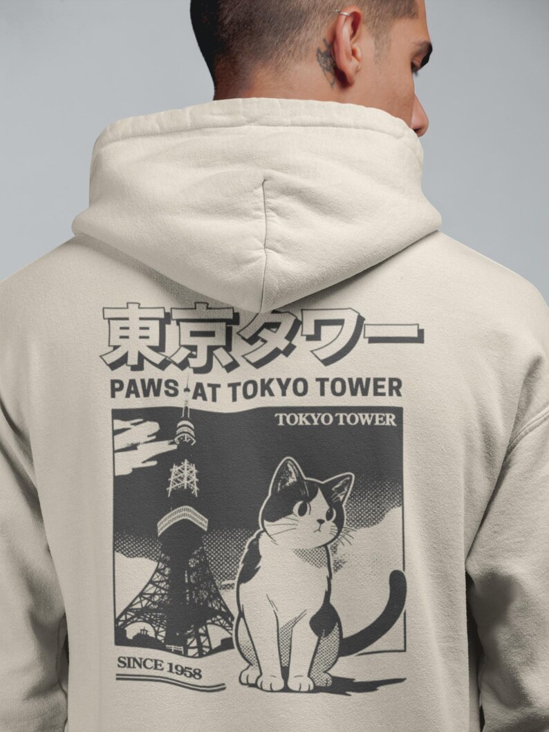 May include: Cream hoodie with a graphic of a cat and the Tokyo Tower. The design includes Japanese text and the phrase "Paws at Tokyo Tower" with "Since 1958" below the image. The cat is black and white.