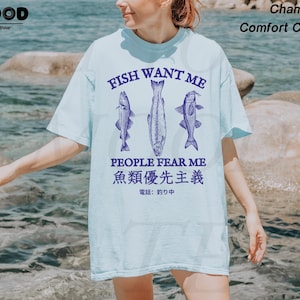 May include: Light blue t-shirt with the text "FISH WANT ME PEOPLE FEAR ME" and Japanese text. The shirt features three fish illustrations in a dark blue color. The shirt is made of a soft, comfortable material.