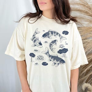 May include: Cream-colored t-shirt featuring a detailed illustration of koi fish swimming among lily pads and flowers. The fish are in shades of blue and white, creating a serene aquatic scene. The shirt has a relaxed fit.