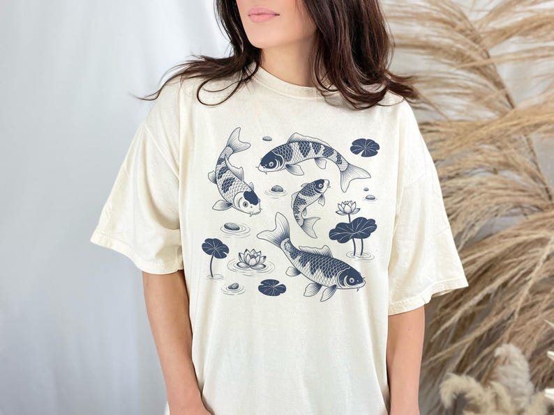May include: Cream-colored t-shirt featuring a detailed illustration of koi fish swimming among lily pads and flowers. The fish are in shades of blue and white, creating a serene aquatic scene. The shirt has a relaxed fit.