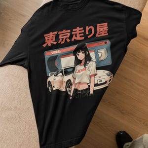 Op de afbeelding: Zwart T-shirt met een witte auto en anime-meisje graphic. De graphic bevat de tekst "Tokyo Drift" en "JDM".