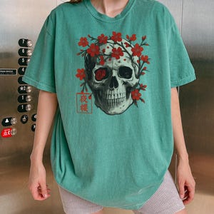 May include: Teal-colored t-shirt featuring a skull graphic adorned with red flowers. The skull has one red eye and is surrounded by a floral wreath. The Japanese characters "夜桜" are printed below the skull.