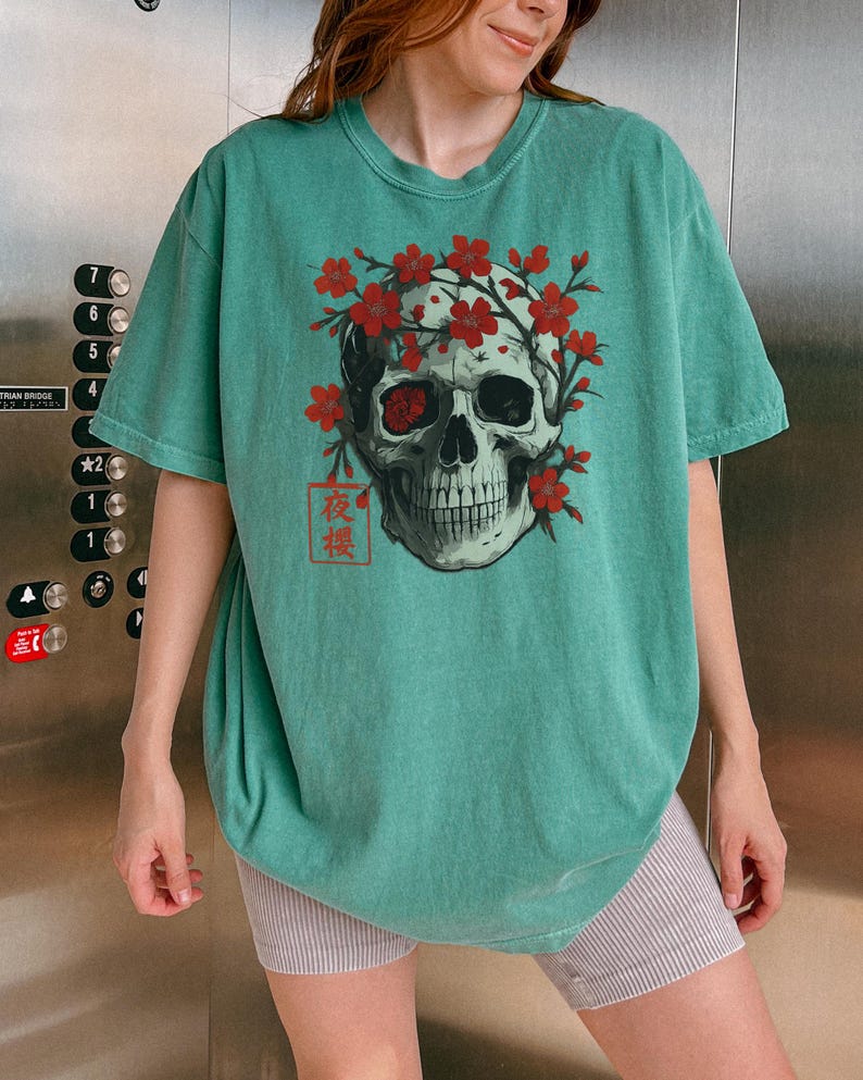 May include: Teal-colored t-shirt featuring a skull graphic adorned with red flowers. The skull has one red eye and is surrounded by a floral wreath. The Japanese characters "夜桜" are printed below the skull.
