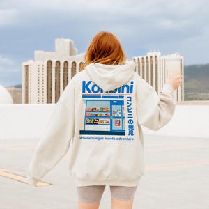 May include: A light gray hoodie with a graphic of a Japanese convenience store on the back. The design includes the word "Konbini" in blue and white, along with Japanese text and the phrase "Where hunger meets adventure."