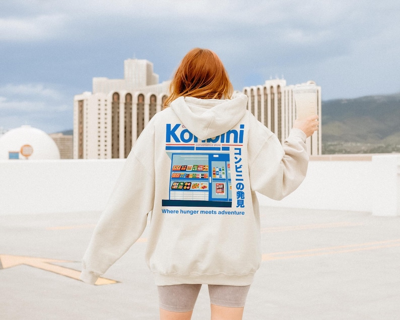 May include: A light gray hoodie with a graphic of a Japanese convenience store on the back. The design includes the word "Konbini" in blue and white, along with Japanese text and the phrase "Where hunger meets adventure."