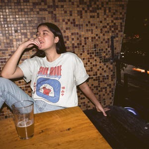 May include: A light-colored t-shirt with the words "SOUL MODE" in red and "just existing" in blue. The shirt features a graphic of a sushi roll. The person is sitting at a wooden table with a glass of beer.