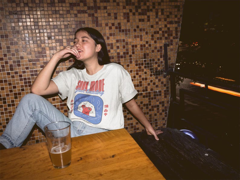 May include: A light-colored t-shirt with the words "SOUL MODE" in red and "just existing" in blue. The shirt features a graphic of a sushi roll. The person is sitting at a wooden table with a glass of beer.