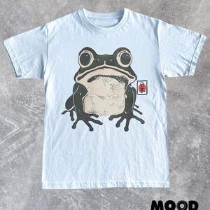 May include: Light blue t-shirt featuring a large graphic of a frog with big eyes and a light beige belly. A small Japanese symbol is on the right side of the frog. The text "MOOD Set Your" is at the bottom.