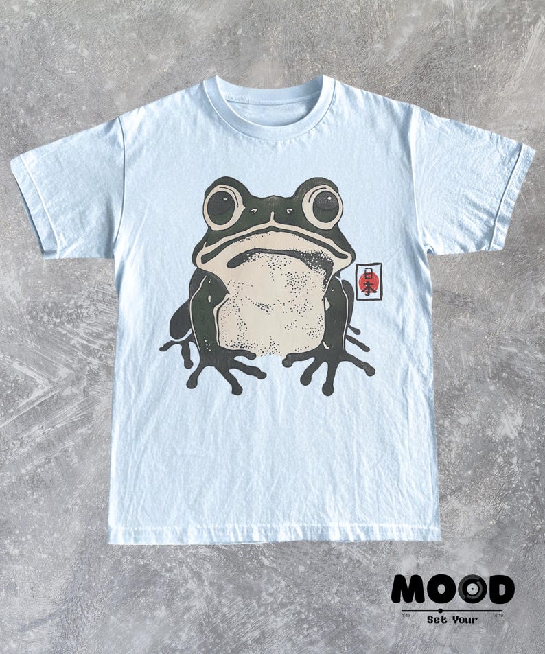 May include: Light blue t-shirt featuring a large graphic of a frog with big eyes and a light beige belly. A small Japanese symbol is on the right side of the frog. The text "MOOD Set Your" is at the bottom.