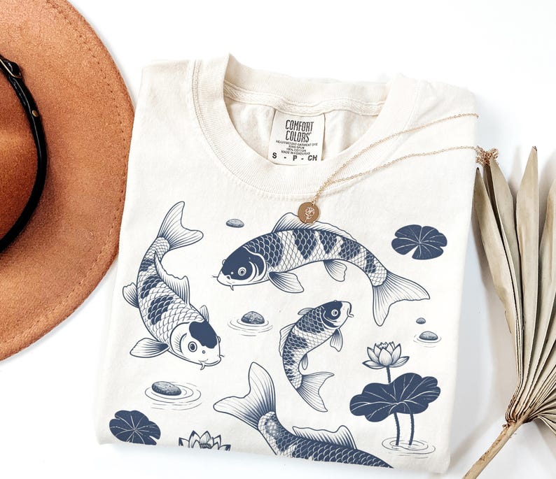 May include: Cream-colored t-shirt featuring a navy blue illustration of koi fish swimming among lily pads and stones. A gold necklace with a pendant rests on the shirt. The shirt has a Comfort Colors tag.